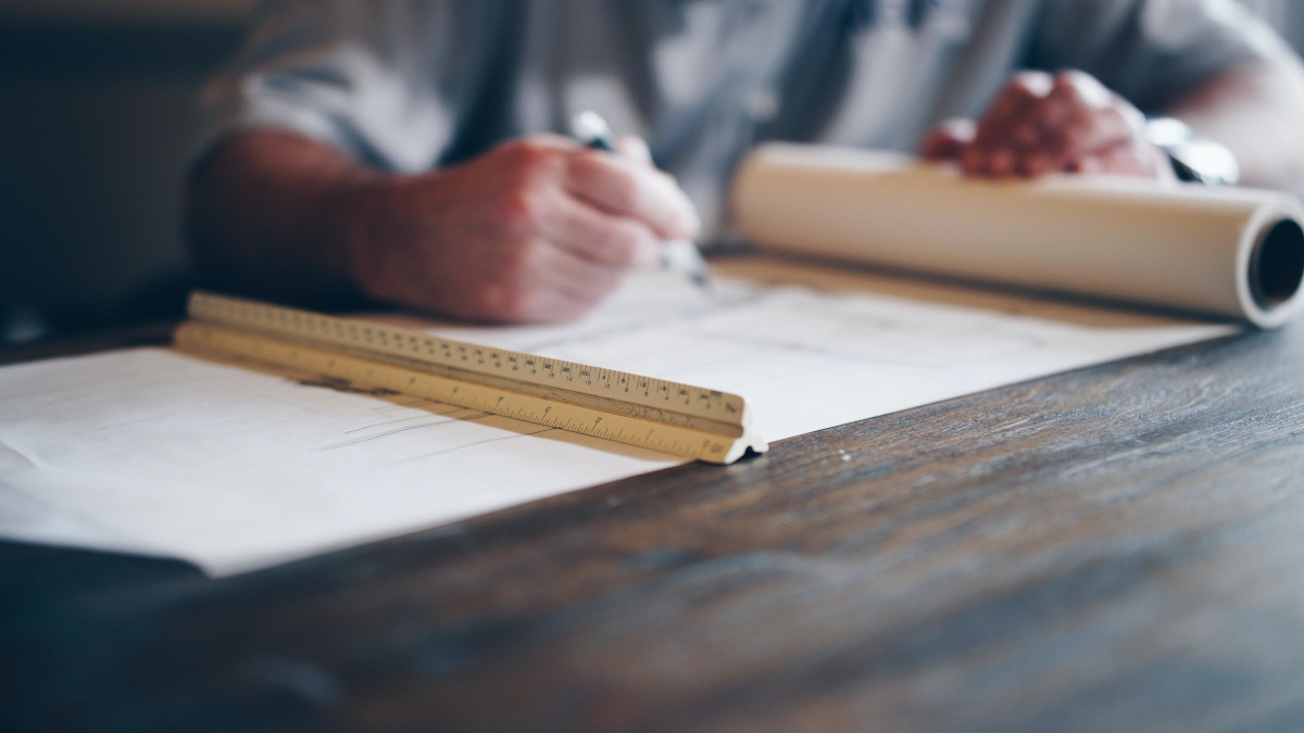 Escritorio con un plano y una regla encima. Al fondo se ve la mano difuminada de un arquitecto, técnico o aparejador que sostiene una pluma